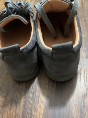 Mörkgrå sneakers med nitar - Säljer ett par mörkgrå sneakers från Christian Louboutin med coola nitar på tån. Skorna har snörning och är gjorda i mocka med en platt sula och rund tå. Insidan är ljusbeige och skorna har en unik edgy look som sticker ut.