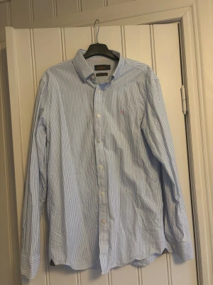 Blårandig skjorta från Morris Stockholm - Säljer en klassisk blårandig skjorta från Morris Stockholm i storlek M. Skjortan har långa ärmar, vit knappslå och diskret logga broderad på bröstet. Tillverkad i mjuk bomull med snygg krage och tidlös design.