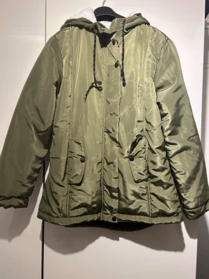 Helt ny! Olivgrön teddyfodrad parkajacka - Helt ny! Olivgrön parkajacka med mjukt vitt teddyfoder, huva med snörning och praktiska fickor med knappar. Jackan har dragkedja och tryckknappar framtill, samt elastisk midja bak för skön passform. Perfekt för kalla dagar med sin fluffiga insida.