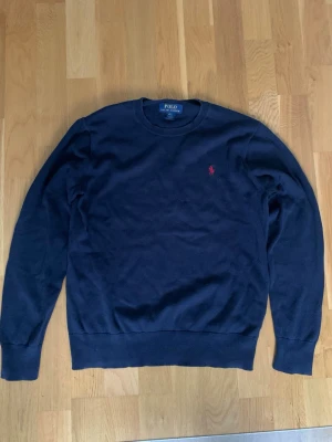 Marinblå tröja från Polo Ralph Lauren - Säljer en marinblå tröja från Polo Ralph Lauren med klassisk röd logga på bröstet och snygga armbågslappar i mörkare tyg. Tröjan har rund halsringning och långa ärmar, perfekt för dig som gillar stilrena och tidlösa plagg. 