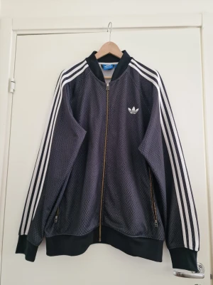 Adidas Originals träningsjacka 2XL - Snygg Adidas Originals herr träningsjacka i mörkgrå/svart med diskret diamantmönster och klassiska vita tre ränder längs ärmarna. Ikoniskt Adidas-Trefoil-tryck på bröstet och dragkedja i guld-look som ger en riktigt cool retrostil.  Storlek: 2XL Perfekt skick  En modell som inte längre finns i butik och kan vara svår att få tag på – perfekt både för samlare och för dig som vill sticka ut med en unik Adidas-look!