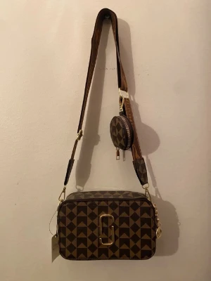 Brun rutig axelväska Jingpindaishu - Snygg brun axelväska med rutigt mönster och guldfärgade detaljer. Väskan har en bred axelrem med text och motiv, samt en liten rund extraväska med dragkedja. Materialet är syntet och väskan har flera guldfärgade dragkedjor och hängen.