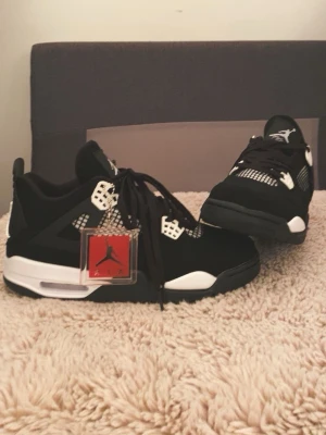 Svarta Nike Air Jordan 4 sneakers - Säljer ett par svarta Nike Air Jordan 4 sneakers med vita detaljer och meshpaneler. Skorna har den klassiska Jumpman-loggan på plösen och hälen, samt snörning och en chunky sula. Materialet är en mix av mocka och syntet. Riktigt snygga och ikoniska sneakers för dig som gillar streetwear.