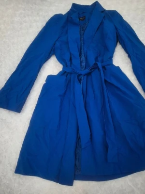 Blå trenchcoat från Bialcon, strl 40 - Snygg blå trenchcoat från Bialcon med klassisk krage och knytbälte i midjan. Jackan har långa ärmar och praktiska fickor på sidorna. Materialet är lätt och har en slät finish, perfekt för dig som gillar stilrena och färgstarka plagg.Den har några små vita fläckar som syns på sista bilden. Den är en liten storlek; jag tror att den skulle passa en storlek S.💙🛍