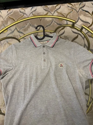 Grå Moncler pikétröja - Säljer en stilren grå pikétröja från Moncler med klassisk krage och knappslå. Tröjan har röda och vita ränder på krage och ärmslut samt Moncler-logga broderad på bröstet. Materialet är mjuk bomull och modellen är normal passform med korta ärmar.