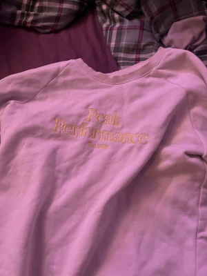 Ljuslila Peak Performance sweatshirt - Säljer en ljuslila sweatshirt från Peak Performance med broderad logga i gult framtill. Tröjan har rund hals, ribbade muddar och är långärmad. Perfekt för dig som vill ha en chill och snygg vardagslook.