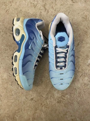 Nike Air Max Plus blå sneakers - Nike Air Max Plus sneakers i ljusblå och marinblå färg med vita meshpaneler och vita snören. Snygg vågig design på ovansidan, synliga Air Max-enheter i sulan och Foot Locker-tag. Skorna har rund tå och platt sula, tillverkade i syntet och mesh.