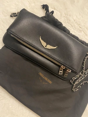 Zadig väska - Snygg svart axelväska i skinn från Zadig & Voltaire med vinge på framsidan. Väskan har två dragkedjor, en framtill och en baktill. Säljer den för att den inte kommer till användning, den är i mycket bra skick och jag har aldrig använt den. Jag köpte den begagnat från Plick.