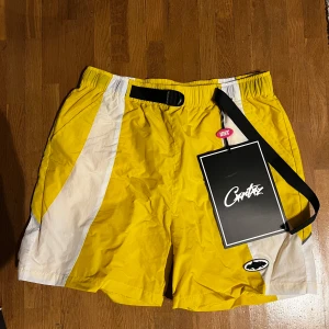 Corteiz shorts - Snygga gula shorts med vita detaljer från Corteiz . De har en elastisk midja med svart bälte och en cool logga på framsidan. Perfekta för en sportig look eller en avslappnad dag.