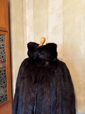 Mörkbrun pälsjacka med krage - Maffig svart pälsjacka med hög krage och långa ärmar. Jackan har en klassisk rak form och stängs med en knapp framtill. Den har även fickor framtill. Den ät i äkta päls och insidan har detaljer i skinn (se bild). Storlek 38/40. Den är flera år gammal därav inget märke på den. Kappan har inga skador eller defekter. Den är väl omhändertagen.