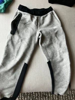 Grå mjukisbyxor från Nike, storlek L junior - Snygga grå mjukisbyxor från Nike med svarta detaljer vid midja, ficka och benslut. Byxorna har en dragkedja på ena fickan och Nike-logga tryckt på framsidan. Perfekta för chill tillverkade i mjukt material för extra komfort.