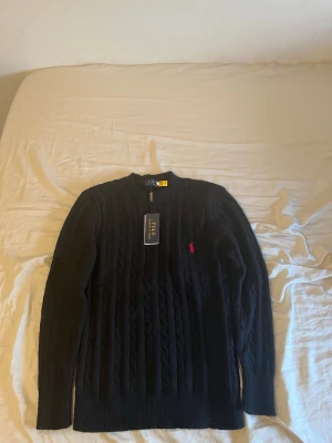 Svart stickad tröja från Polo Ralph Lauren - Svart kabelstickad tröja från Polo Ralph Lauren med rund hals och lång ärm. Tröjan har ett rött broderat Polo-logo på bröstet och ribbade muddar vid hals, ärmslut och nederkant. Perfekt för dig som gillar klassisk och stilren design.