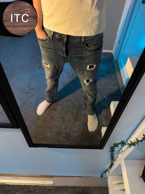 Jeans med slitningar - Säljer ett par blå jeans med snygga slitningar och hål på framsidan av benen. Modellen har tapered fit och klassisk femficksdesign. Jeansen är tillverkade i jeansmaterial och har en mörk tvätt med ljusare detaljer kring slitningarna.
