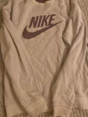Beige sweatshirt från Nike med logga - Säljer en beige sweatshirt från Nike i storlek S. Tröjan har en stor mörklila Nike-logga på bröstet och är långärmad med ribbade muddar vid ärmslut och nederkant. Perfekt för chill dagar och streetwear-stil.