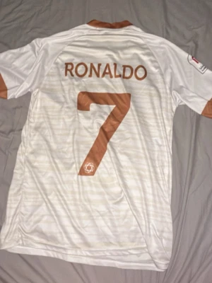 Vit och brun Al Nassr Ronaldo tröja - Säljer en officiell Al Nassr matchtröja med Ronaldo och nummer 7 på ryggen. Tröjan är vit med bruna detaljer på krage och ärmslut, samt brunt tryck fram och bak. Materialet är syntet och har subtila ränder. Perfekt för fotbollsälskare!