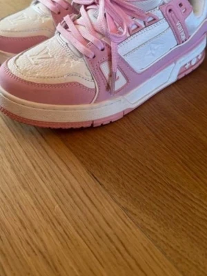 Louis Vuitton sneakers i rosa och vitt - Säljer ett par snygga Louis Vuitton sneakers i rosa och vitt med klassiska LV-detaljer på sidan och sulan. Skorna har snörning, rund tå och platt sula. Materialet är en mix av skinn och syntet, med präglade loggor och mönster på ovandelen. Perfekt för dig som gillar exklusiva sneakers.