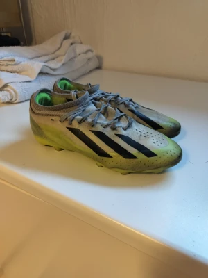 Adidas fotbollsskor grå och lime - Säljer ett par Adidas fotbollsskor med grå ovandel, limegröna detaljer och svarta ränder. Skorna har snörning och platt sula med dobbar för bra grepp på planen. Insidan är limegrön och materialet är syntet. Perfekt för dig som vill ha snygga och funktionella skor till matchen.