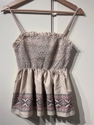 Beige topp - Topp som sitter tajt runt bysten och löst med volanger nedtill. Toppen har justerbara band och ett fint mönster. Toppen är aldrig använd. 