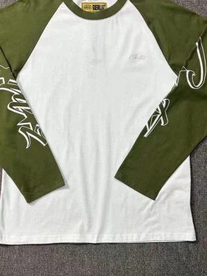 Vit och grön långärmad topp Guerilla - Säljer en cool långärmad topp från Guerilla med vita och olivgröna färgblock. Ärmarna har snygga grafiska tryck i vitt som ger en streetwear-vibe. Toppen har rund hals och är tillverkad i mjuk bomull, perfekt för chill dagar.    Passar folk som har storlek L också 