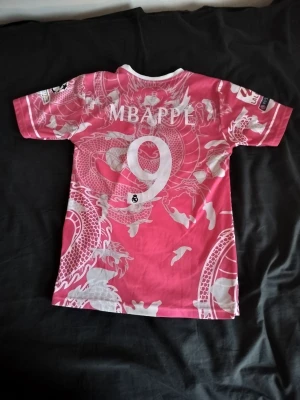 Real Madrid tröja med Mbappe på ryggen - Bilderna visar en rosa Real Madrid fotbollströja med drakmönster och Mbappé-tryck.