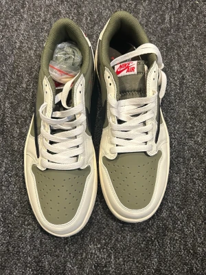 Nike Air Jordan 1 Travis Scott Olive - Nike Air Jordan 1 Travis Scott i olivgrön och vit med svarta detaljer. Skorna har klassisk snörning, rund tå och platt sula. Märket syns tydligt på tungan och hälen, med röd broderad logga och Travis Scott-emblem. Materialet är skinn och syntet, med olivgröna inslag och vit bas.