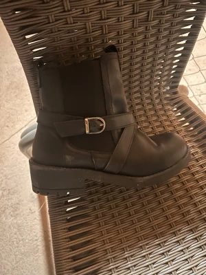 Svarta boots med spänne och dragkedja - Säljer ett par svarta boots i skinn med rund tå och platt sula. Skorna har snygga detaljer med spänne i metall och dragkedja på sidan för enkel på- och avtagning. Perfekt för dig som gillar stilrena och robusta skor med lite edge.