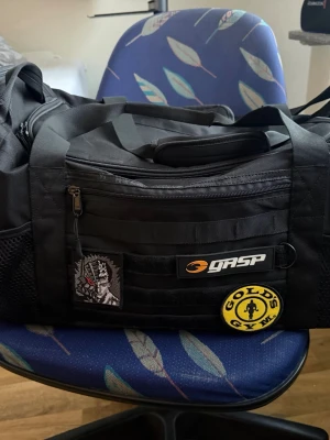 Svart träningsväska från GASP - Stor svart träningsväska från GASP med orange logga och flera fack. Väskan har meshfickor på sidorna, robusta handtag och axelrem med vaddering. Utsmyckad med Gold's Gym-patch och anime-inspirerad patch framtill. Perfekt för gymmet och sport.