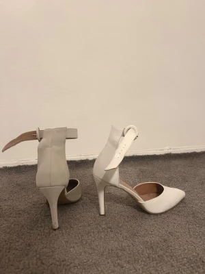 Vita pumps med spetsig tå och klack - Säljer ett par stilrena vita pumps med spetsig tå och hög klack. Skorna har en bred rem runt ankeln med spänne och är tillverkade i skinnimitation. Perfekta för dig som gillar en elegant look och vill ha något som sticker ut.