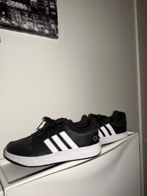 Svarta Adidas sneakers med vita ränder - Säljer ett par klassiska svarta Adidas sneakers med vita ränder på sidorna och vit sula. Skorna har snörning och är tillverkade i syntetmaterial med meshdetaljer vid hälen. Perfekta för dig som gillar stilrena och sportiga skor.