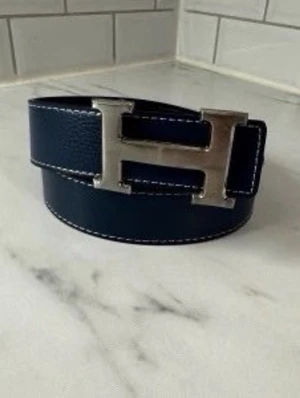 Mörkblå skinnbälte med H-spänne - Snyggt mörkblått bälte med ett stort H-format metallspänne. Bältet är gjort i skinn med vita kontrastsömmar längs kanterna som ger en cool detalj. Perfekt för att lyfta din outfit med en stilren accessoar.