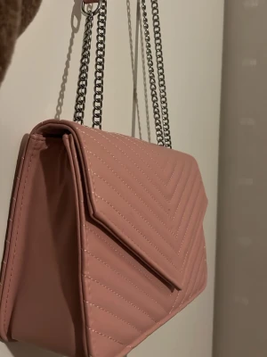 Rosa väska - Säljer en snygg rosa axelväska med silverfärgad kedja. Perfekt för dig som gillar trendiga accessoarer med lite extra detaljer. Själv har jag aldrig använt den, är helt ny✨inget varumärke köptes från en marknad🌺