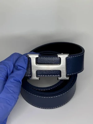 Bälte - Bilden visar ett blått läderbälte med ett stort H-spänne i metall, inte en väst. Ingen information om väst kan ges.