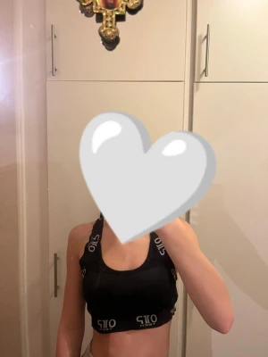 Svart sporttopp - Säljer en svart sporttopp med breda axelband som korsas i ryggen. Toppen har vit logotyptext längs banden och nederkanten. Materialet är stretchigt och passar perfekt för träning. Modellen är croppad och ärmlös med rund hals.