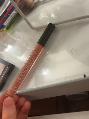 Huda Beauty Lip Gloss Peachy Nude - Säljer ett Huda Beauty Tint Lip Gloss i en snygg peachy nude-nyans. Kommer i en smidig tub med svart lock och transparent hylsa som visar färgen. Perfekt för dig som vill ha glansiga och återfuktade läppar med en hint av färg.