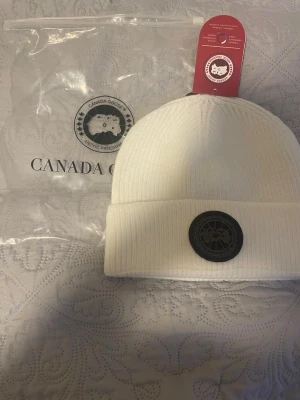 Ingen väst på bilden - Bilden visar en vit mössa från Canada Goose, inte en väst. Mössan är ribbstickad med ett svart märke framtill.