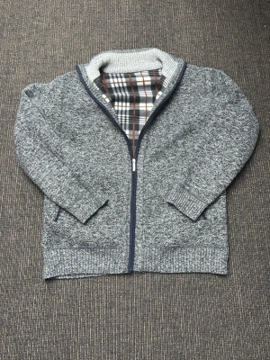 Grå stickad kofta med dragkedja - Säljer en sjukt snygg gråmelerad stickad kofta med dragkedja och högt ribbat krage. Insidan har ett rutigt foder i brunt, vitt och grått som ger extra värme. Koftan har två fickor framtill och är långärmad, perfekt för kyliga dagar. Inga defekter! Hör av er vid minsta fundering!