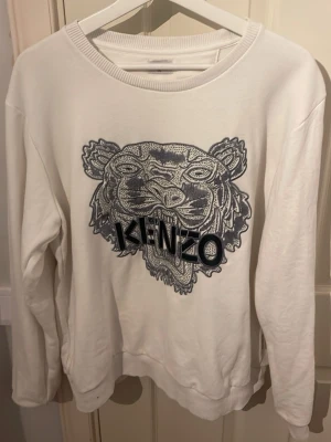 Vit Kenzo sweatshirt med tigertryck - Säljer en vit sweatshirt från Kenzo med det ikoniska tigerbroderiet och loggan i mörkblått framtill. Tröjan har rund hals, långa ärmar och är gjord i mjuk bomull. Perfekt för dig som vill ha en clean men ändå statement look.