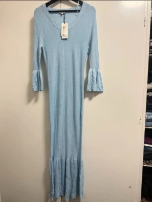 Ljusblå långklänning från Indiska - Supersöt ljusblå långklänning från Indiska med ribbad struktur och volangdetaljer vid ärmslut och nederkant. Klänningen har trekvartsärm och en rundad halsringning. St S 