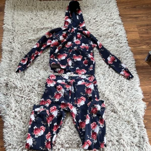 Blommig sett från Cubus - Säljer en mörkblå hoodie och mjukisbyxor från Cubus, tillverkad i ekologisk bomull.  De har ett blommigt mönster i rött, beige och vitt, hoodien har dragkedja framtill och snörning vid huvan.  Perfekt för dig som gillar färgstarka och mjuka plagg. De är i använt skick. Underdelen är i storlek XL och överdelen är i storlek L