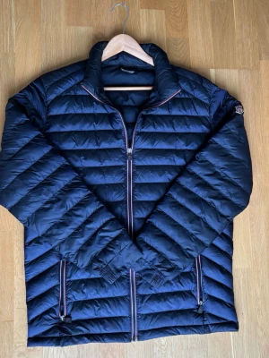 Mörkblå Moncler pufferjacka - Snygg mörkblå pufferjacka från Moncler med horisontella quiltade paneler och coola detaljer som röd, vit och blå dragkedja. Jackan har hög krage, två sidofickor med dragkedja och ett unikt serietryck på insidan. Perfekt för dig som vill ha både stil och värme. 