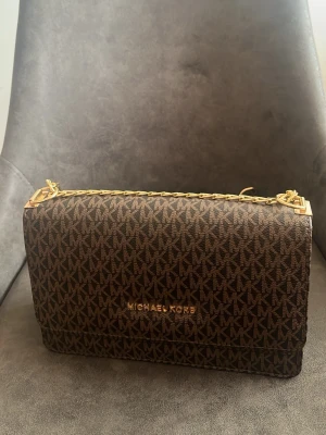 Brun axelväska från Michael Kors - Snygg brun axelväska från Michael Kors med klassiskt MK-mönster över hela väskan. Guldiga detaljer och kedjerem ger en lyxig känsla. Väskan har magnetlås och ett rymligt fack med innerficka som stängs med dragkedja. Perfekt storlek för det viktigaste.