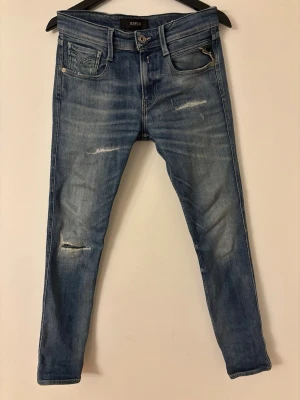 Replay Anbass blå jeans, stl 29 - Säljer ett par Replay Anbass jeans i klassisk blå tvätt med slitningar och små hål för en cool look. Modellen har fem fickor, normal passform och raka ben. Jeansen är gjorda i mjukt bomullsmaterial med snygga detaljer och läderpatch bak i midjan.