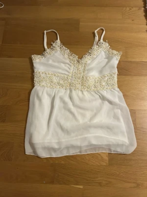 Vit spetsdetaljerad linnetopp från Vila - Säljer en vit linnetopp från Vila i storlek XS. Toppen har tunna axelband, spetsdetaljer med pärlor och paljetter längs halsringning och midja, samt omlottband baktill. Materialet är lätt och luftigt, perfekt för varma sommar dagar. Säljer då den inte används längre💕 (har små fläckar som bara syns på insidan)