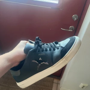 Svarta sneakers från Axel Arigato - Svarta sneakers från Axel Arigato med broderad fågel på sidan och vit platt sula. Skorna har snörning och är tillverkade i skinn med detaljer i mocka vid tån. Klassisk låg modell med stilren design och diskret logga i guld på sidan.