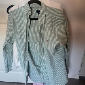 Grönrandig skjorta från Polo Ralph Lauren - Snygg grön skjorta med vita ränder från Polo Ralph Lauren. Klassisk button-down krage, broderad logga på bröstet och knappar framtill. Långärmad och tillverkad i mjuk bomull som känns skön mot huden. Perfekt för dig som gillar stilrena och fräscha plagg.