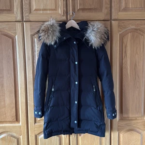 Svart dunjacka med päls från Hollies - Snygg svart dunjacka från Hollies med fluffig naturfärgad päls på huvan. Jackan har långa ärmar, dragkedjor på fickorna och tryckknappar vid ärmslut. Perfekt för kalla dagar och har en klassisk, stilren look.