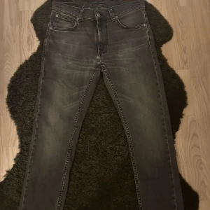 Mörkgrå jeans från Nudie - Snygga mörkgrå jeans från Nudie med klassisk femficksdesign och diskreta slitningar. Jeansen har raka ben och normal passform, med Nudie-loggan broderad på bakfickorna. Perfekta för dig som gillar en stilren look. Jeansen har en liten defekt allra löngst bed på byxbenet som knappt syns. W30/L30 men kan passa längre i längd