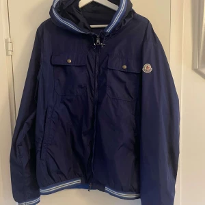 Blå vindjacka från Moncler.  - Vindjacka från Moncler. Jackan är använt i ett år men bara använt några gånger. Perfekt höst och vår jacka. Den är i storlek 5 vilket motsvarar L men passar om man är lång. 185+ så sitter den perfekt.  Skick är väldigt bra. Skriv för mer information. 