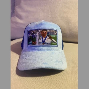 Blå meshkeps Alento med fototryck - CR7 Limited "Five-Time Champion"   Kolla hit alla fotbollsentusiaster! Legendariske Cristiano Ronaldo vid det ikoniska ögonblick när han stolt höll Champions League-trofén högt, med sin hand som graciöst visar alla fem fingrar – en symbol för att han är femfaldig mästare. Begränsad upplaga: Endast 50 stycken