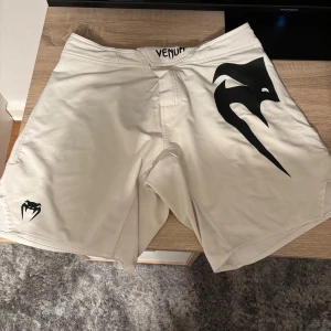 Vita Venum fightshorts i storlek L - Säljer ett par vita Venum fightshorts med svart logga och tryck på både fram- och baksidan. Shortsen har en loose passform och är tillverkade i lätt syntetmaterial, perfekta för träning eller kampsport. Stängs med kardborre och har cool design. Har bara använt dem 5-10 gånger då de var för stora för mig. Köpta för 700 säljer för 400.